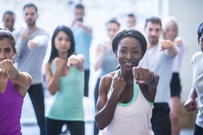 A multi-ethnic group of adults are indoors in a fitness center. They are wearing casual exercise clothing. They are doing a punching motion during an exercise class. A woman of African descent is leading the class.